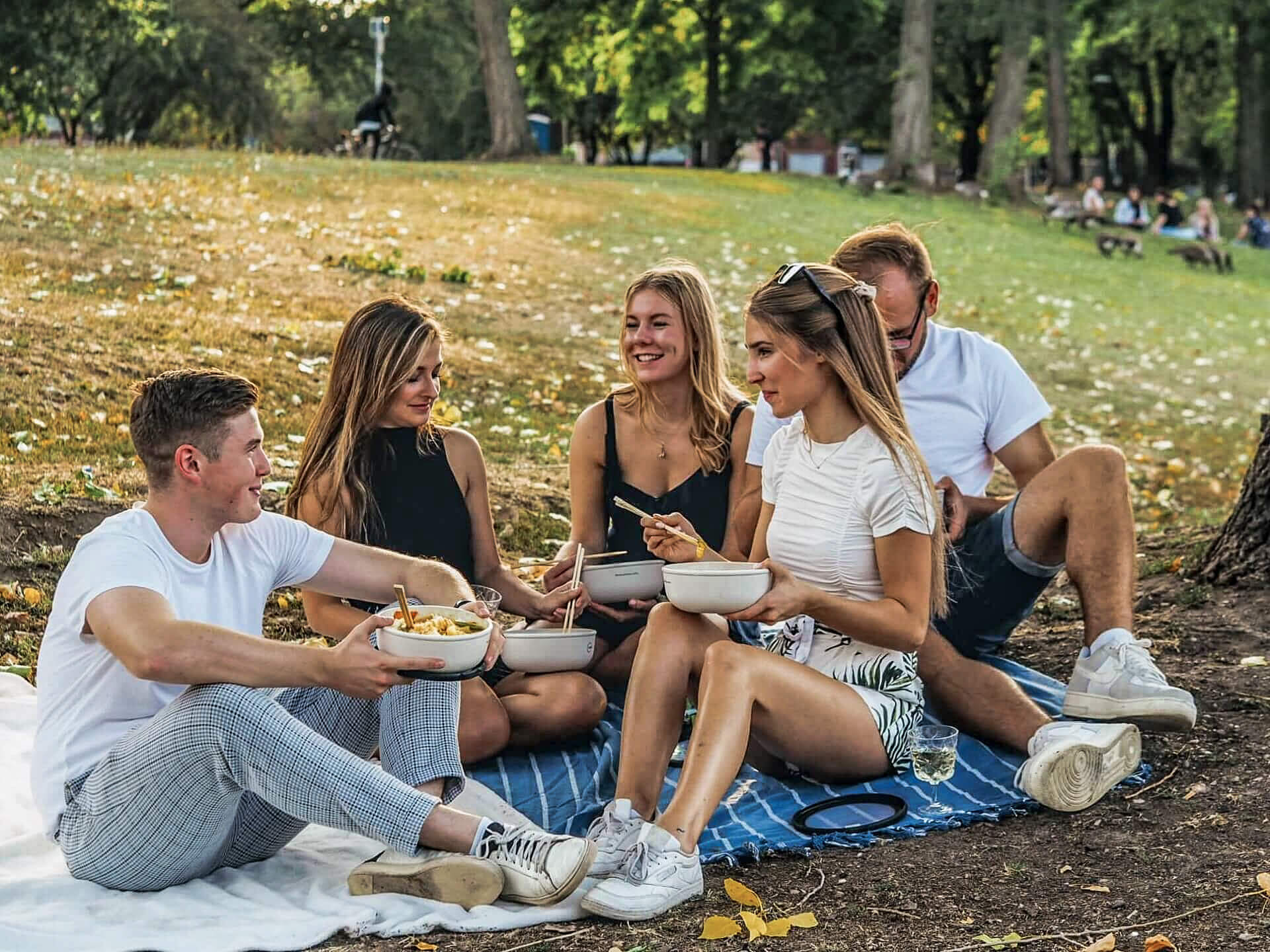 eine junge Gruppe von Menschen sitzt im Park und isst Gerichte aus wiederverwendbaren Behältern
