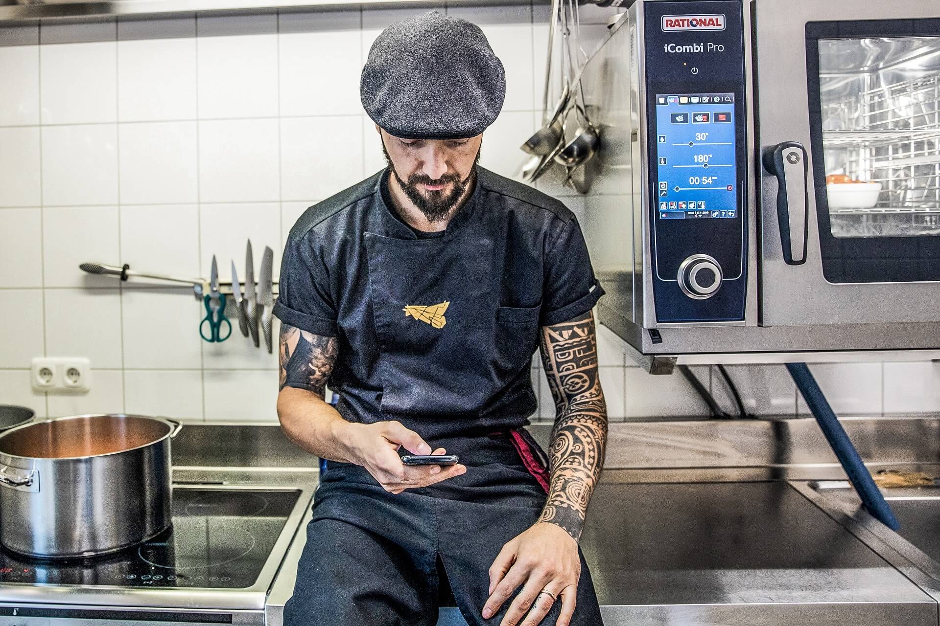Tätowierter Koch sitzt in der Küche und sucht in den sozialen Medien nach Inspirationen und Informationen.