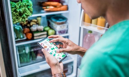 Mit Obst und Gemüse gefüllter Kühlschrank, davor ein Mann mit einem Smartphone, der nach neuem Gemüse sucht