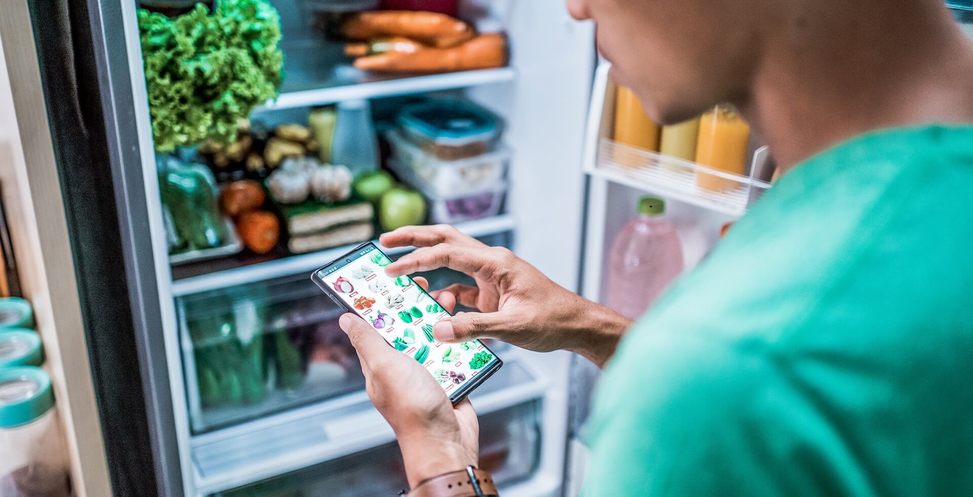 Refrigerator filled with fruits and vegetables, in front of it a man with a smartphone looking for new vegetables