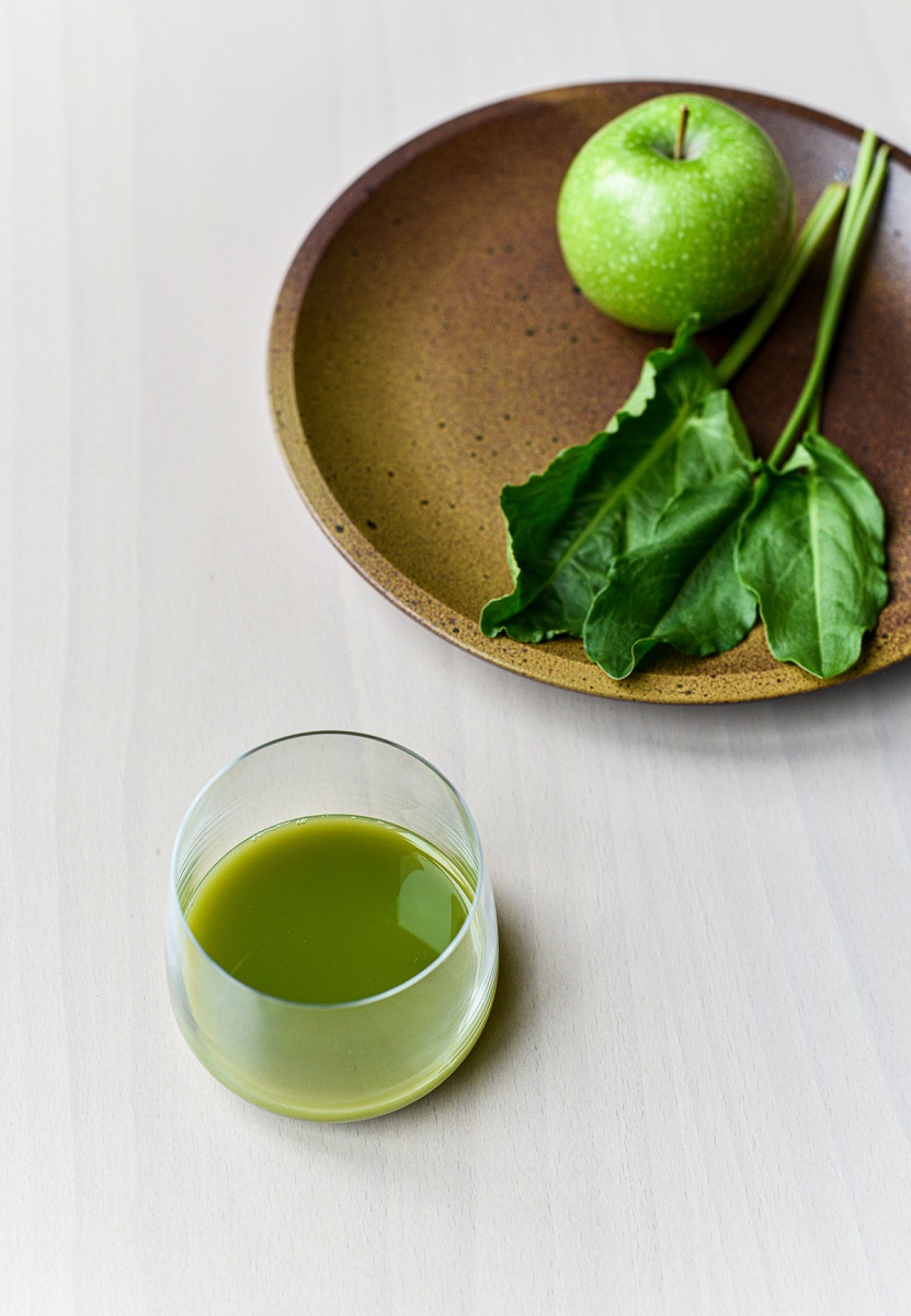An apple on a plate with a glass of green juice next to it