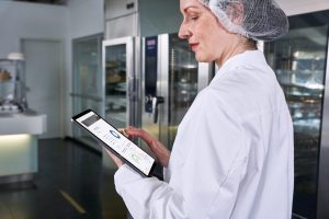 Eine Frau mit Haarnetz steht in einer professionellen Küche und hält ein Tablett mit HACCP Daten