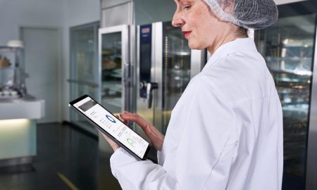 A woman with a hairnet stands in a professional kitchen and holds a tray with HACCP data